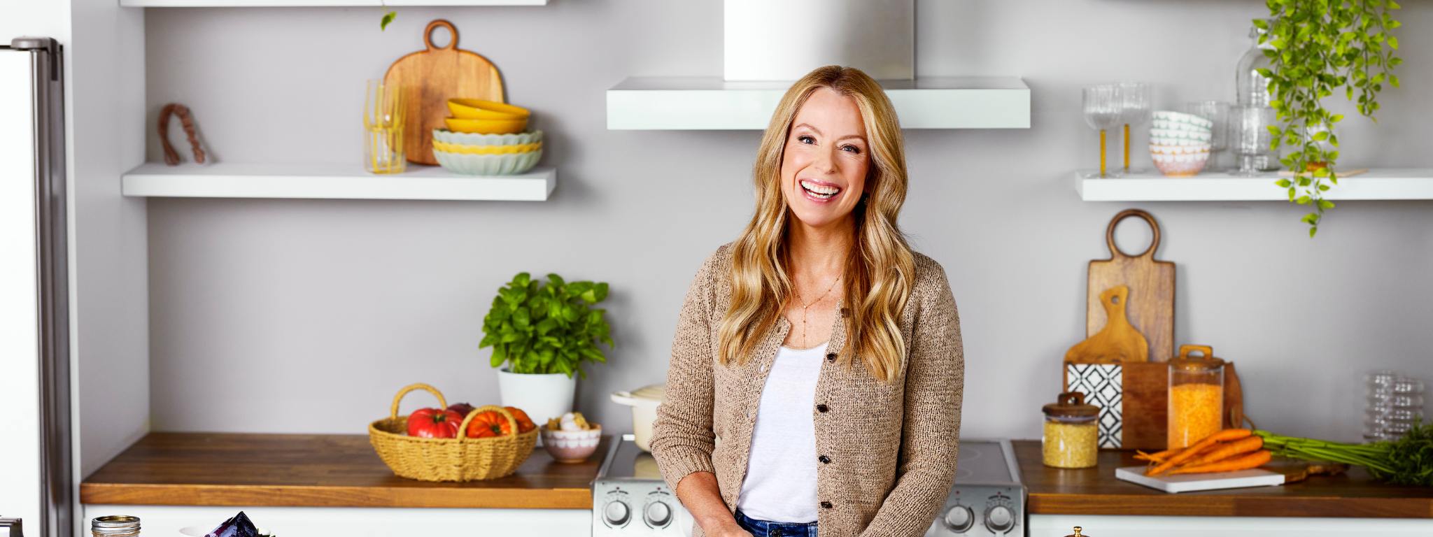 Isabelle Huot, debout dans une cuisine moderne avec des légumes frais, des herbes et des produits de cuisine visibles sur les étagères. Elle coupe des légumes dans ses mains et porte un cardigan beige.