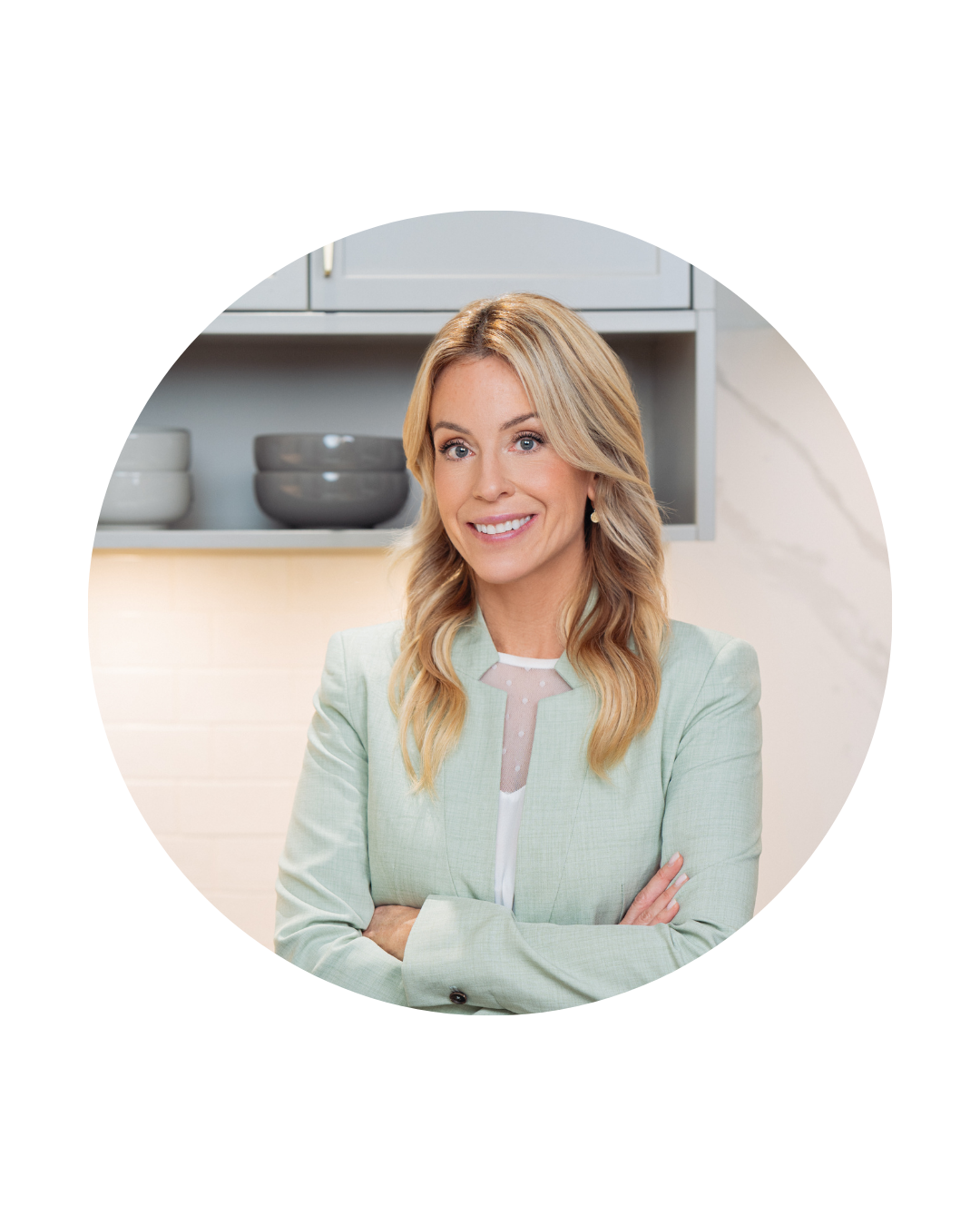 Portrait d’Isabelle Huot souriante, bras croisés, vêtue d’un veston vert pâle, dans une cuisine moderne. Portrait of Isabelle Huot smiling, arms crossed, wearing a light green jacket, in a modern kitchen.