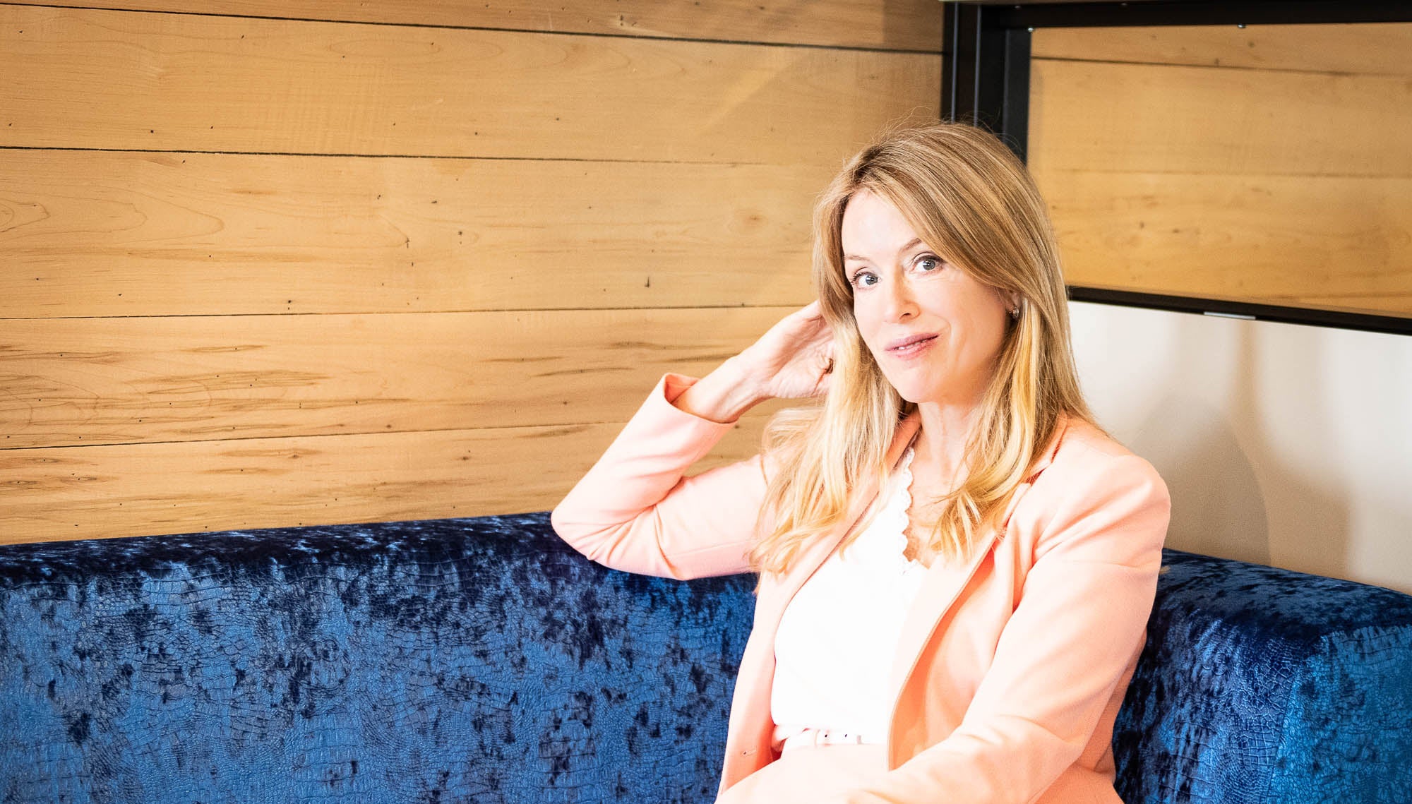 Isabelle Huot assise sur un canapé bleu, vêtue d’un veston pêche, dans un décor intérieur moderne avec mur en bois clair. Isabelle Huot sitting on a blue sofa, wearing a peach jacket, in a modern interior setting with light wood walls.