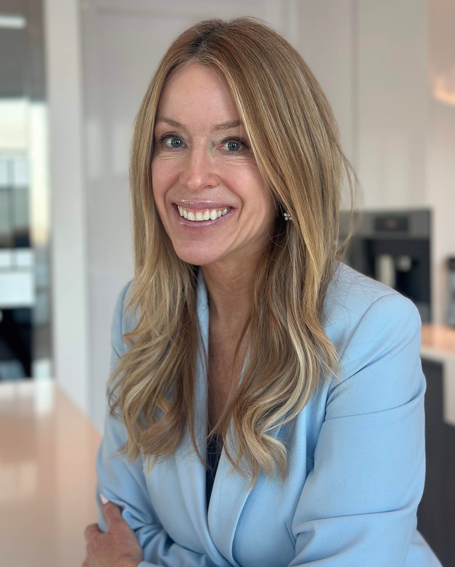 Portrait d’Isabelle Huot souriante, vêtue d’un veston bleu pâle, dans un environnement intérieur lumineux et moderne. Portrait of Isabelle Huot smiling, wearing a light blue jacket, in a bright and modern indoor setting.
