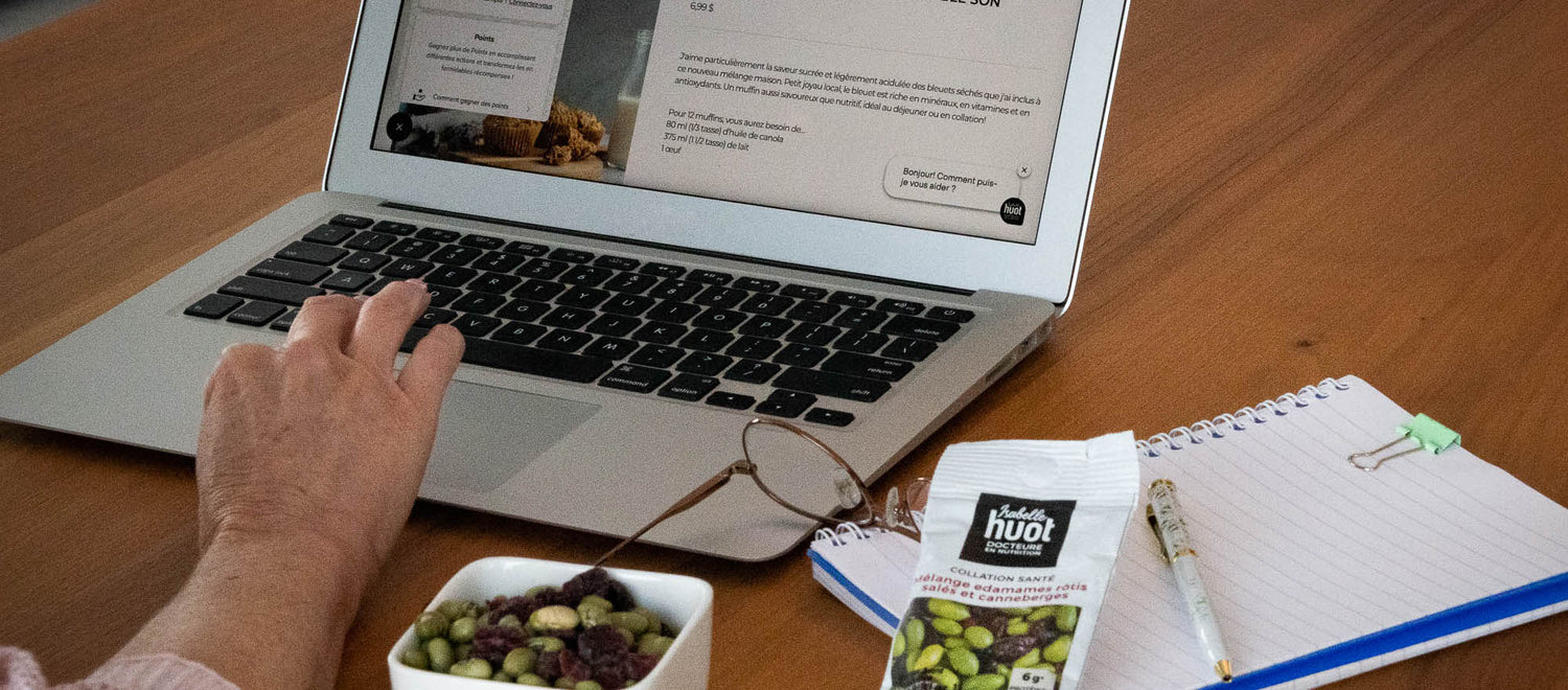 Main utilisant un ordinateur portable sur une table en bois, avec carnet de notes, lunettes, collation santé Isabelle Huot et bol d’edamames et canneberges. Hand using a laptop on a wooden table, with notebook, glasses, healthy snack Isabelle Huot, and bowl of edamame and cranberries.